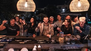 A group of people at a bar with wicker pendant lights, a bar counter with various bottles and glasses, and a smartphone on the counter. - qema Arequipa (Arequipa)