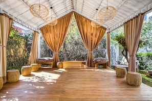A covered outdoor seating area with wooden flooring, woven poufs, and a bench.