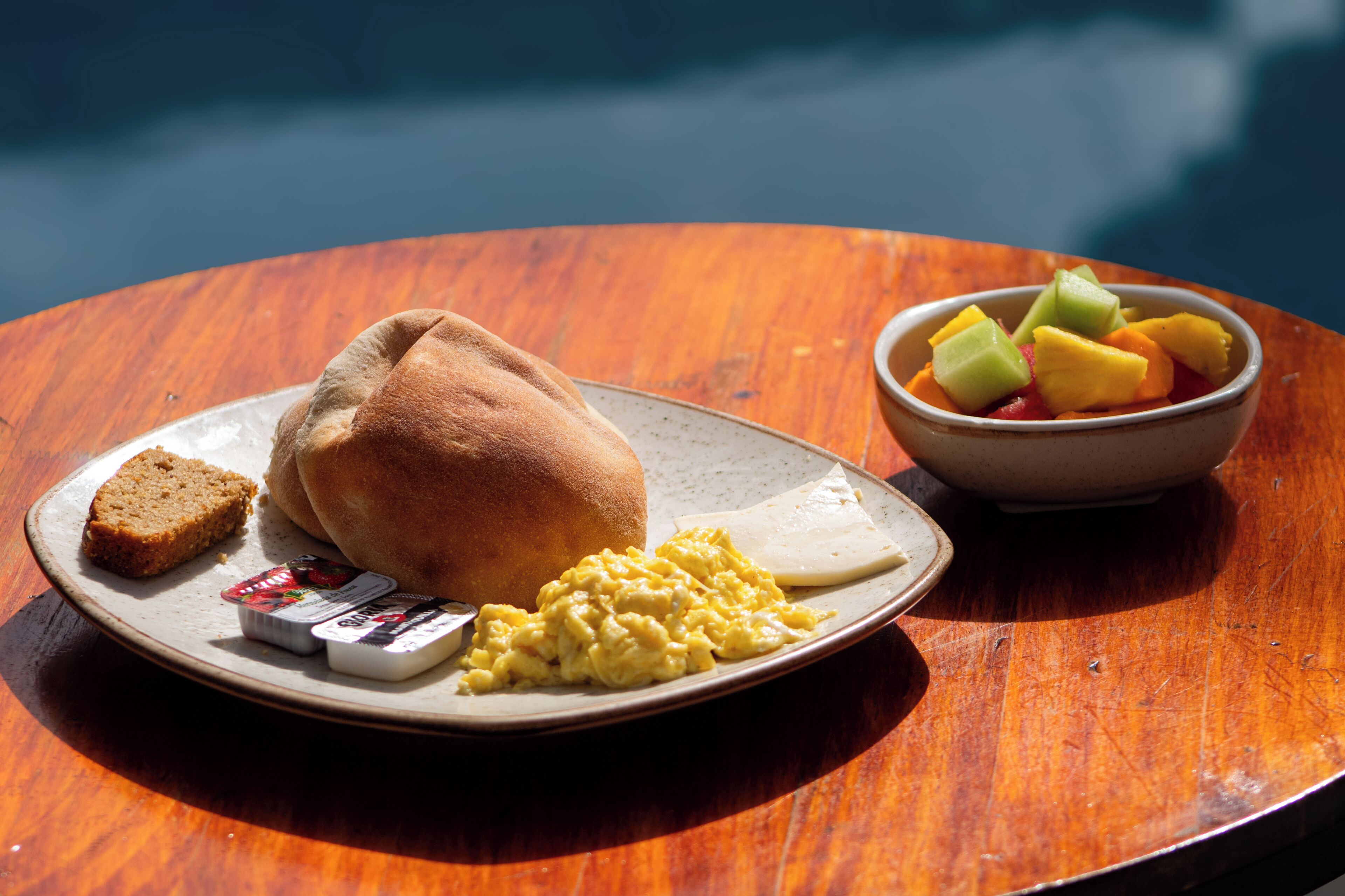 A breakfast plate with scrambled eggs, toast, and a bowl of fruit on a wooden table.