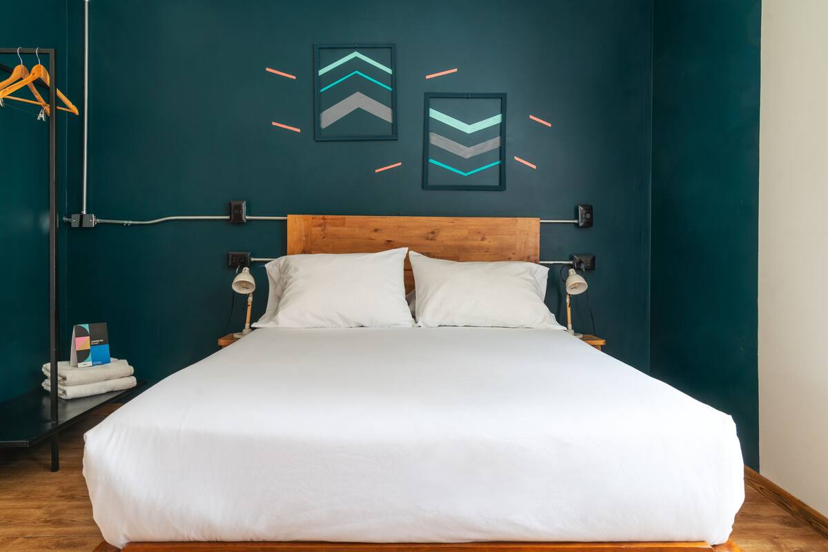 Una cama bien hecha en una habitación con paredes verde azulado, dos cuadros enmarcados y un cabecero de madera.