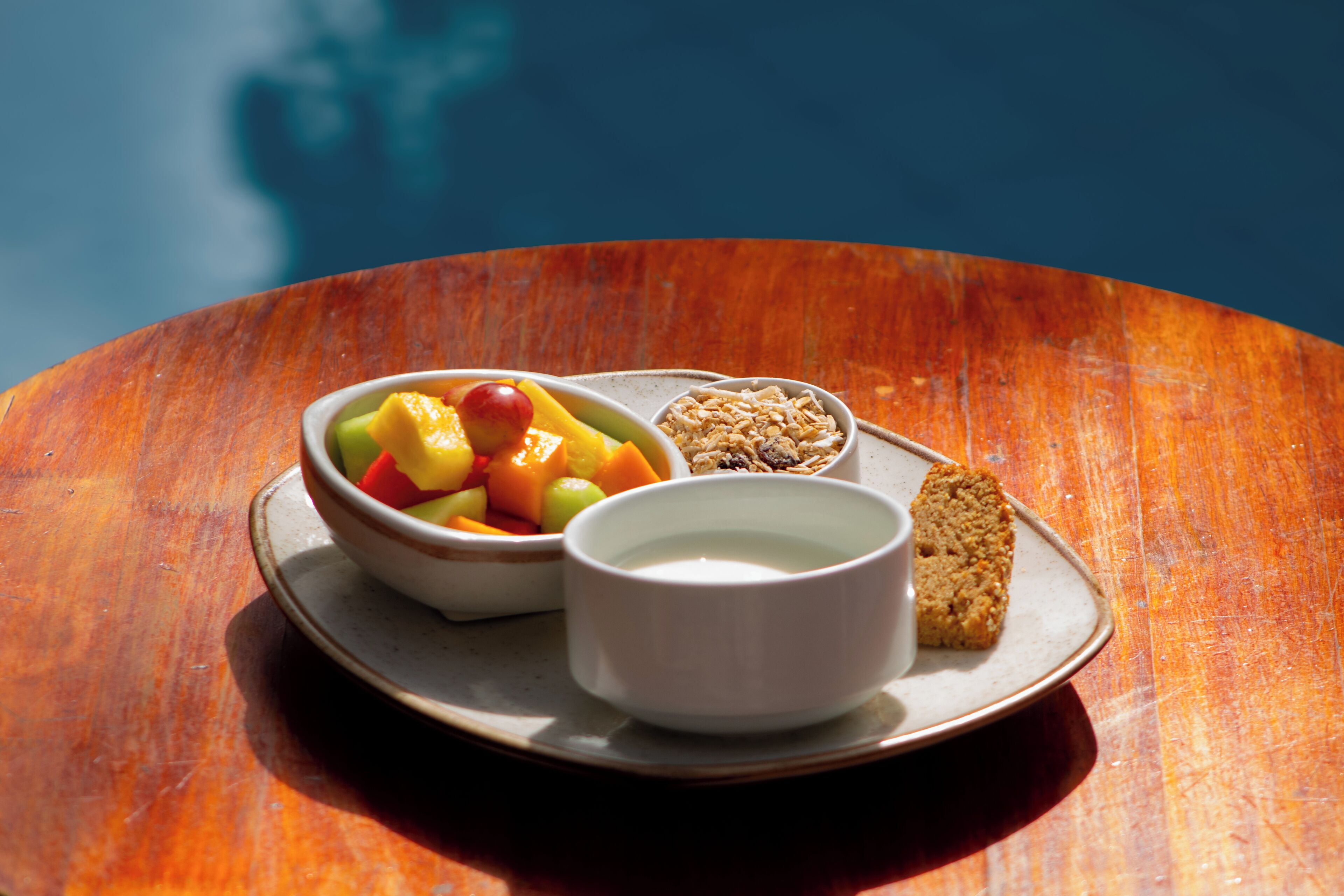 Un plato con un cuenco de fruta, un tazón de cereal, una taza de leche y una rebanada de pan sobre una mesa de madera.