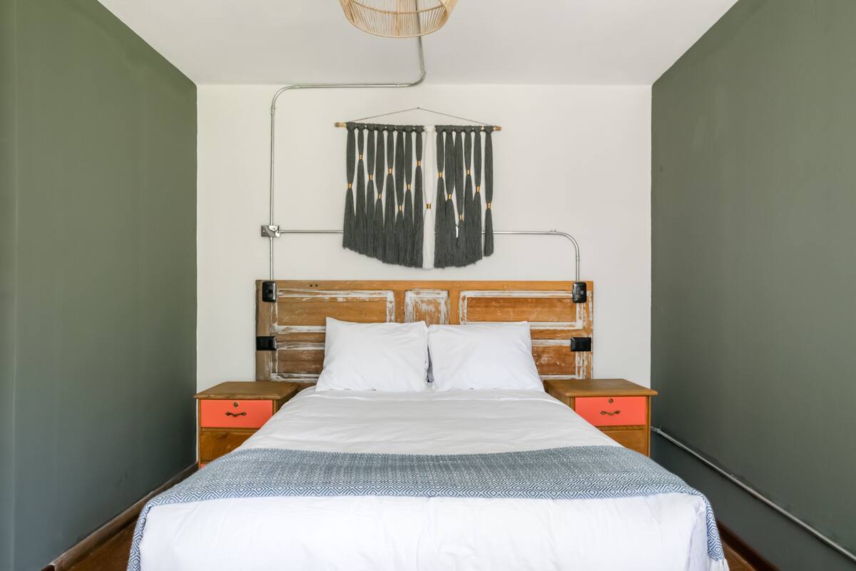 Una cama bien hecha con sábanas blancas, un cabecero de madera y una cortina de macramé colgante. Hay dos mesitas de noche de madera con cajones, una de ellas naranja.