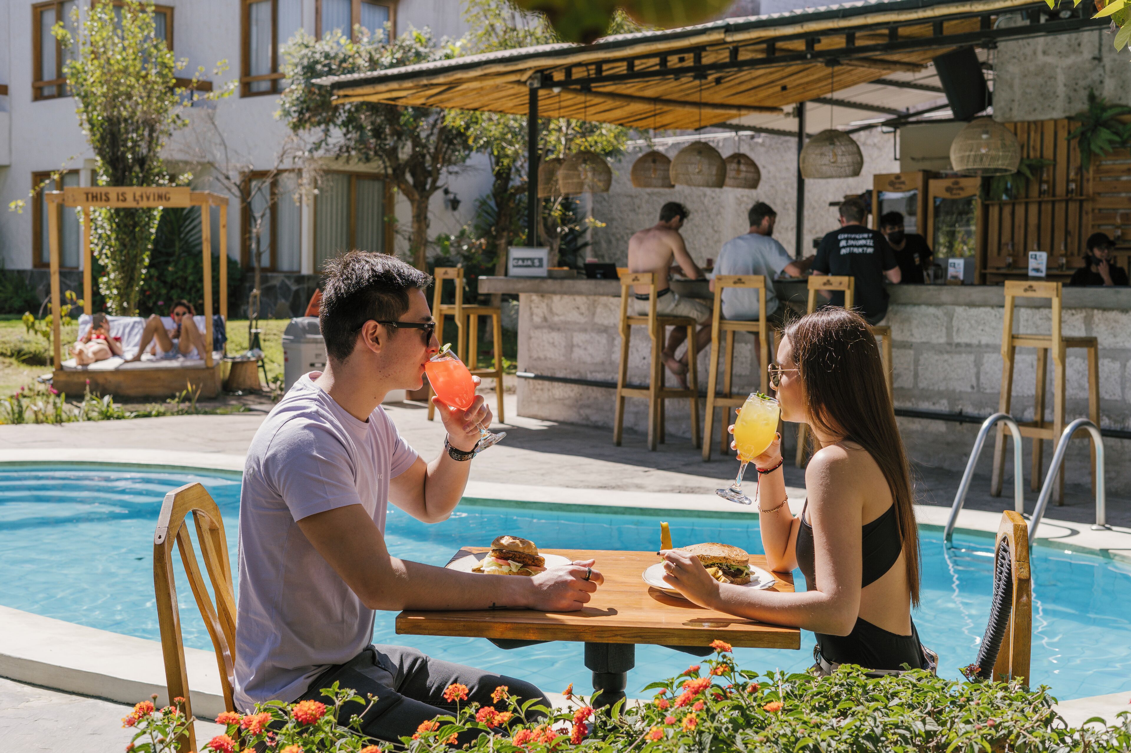 A poolside dining area with wooden chairs and tables, a pool, and a bar area in the background.