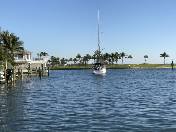 Un velero en aguas tranquilas, amarrado cerca de una casa con un muelle y palmeras al fondo.