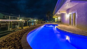 A nighttime swimming pool with a blue-lit edge, surrounded by a pebble-lined deck and a building with a purple facade. - Salty Anglers: Waterfront Heated Pool, Dock & Walk to Sombrero Beach in Marathon (Marathon)