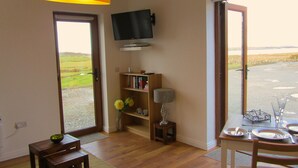 A room with a wooden floor, a bookshelf, a television mounted on the wall, and a dining area with a table set for four. - Atlantic Apartment. Wonderful views of the Wild Atlantic Ocean and mountains (Renvyle)
