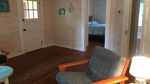 A room with wooden floors, a chair, a bed with a floral bedspread, and a door leading to another room. - Short walk to beach or Pier Village, We consider FLETC (Saint Simons Island)