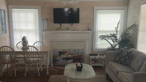 A living room with a ceiling fan, a flat-screen TV mounted above a fireplace, a sofa, a coffee table, and a dining area with chairs. - Short walk to beach or Pier Village, We consider FLETC (Saint Simons Island)