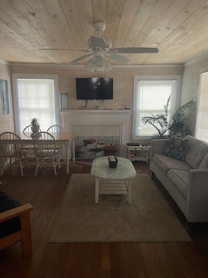 Cozy living room with a high ceiling, wooden beams, and a fireplace. Features a sofa, coffee table, dining table, and a flat-screen TV.