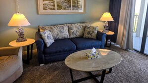 A living room with a blue sofa, two lamps, a framed artwork, and a coffee table. - OCEAN FRONT Classy, Beachy Double Balcony Condo #306 at Cape Winds Resort (Cape Canaveral)