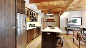 Eine moderne Küche mit Holzausstattung, einem Edelstahlkühlschrank, einer zentralen Kochinsel mit Hockern, einem Essbereich mit Tisch und Stühlen sowie einem an der Wand montierten Fernseher.