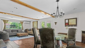 A spacious living room with a high ceiling, wooden beams, a chandelier, and a large window with a view of greenery.