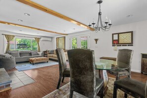 A spacious living room with a high ceiling, wooden beams, a chandelier, and a large window with a view of greenery.