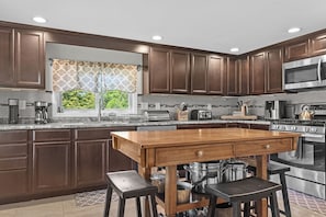 A modern kitchen with dark wood cabinets, stainless steel appliances, a large island with stools, and a window with patterned curtains.