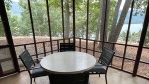 Mesa redonda con cuatro sillas en un espacio exterior cubierto, con vista a árboles y un cuerpo de agua.