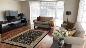 A living room with a sofa, a coffee table, a TV on a stand, and a large window with blinds.