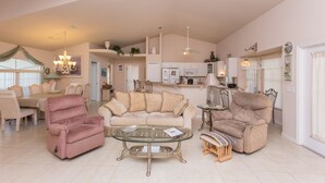 Un vaste espace de vie comprenant un canapé beige, un fauteuil inclinable rose et une table basse en verre. On y trouve également un coin repas avec une table et des chaises, ainsi qu’une cuisine à l’arrière-plan.