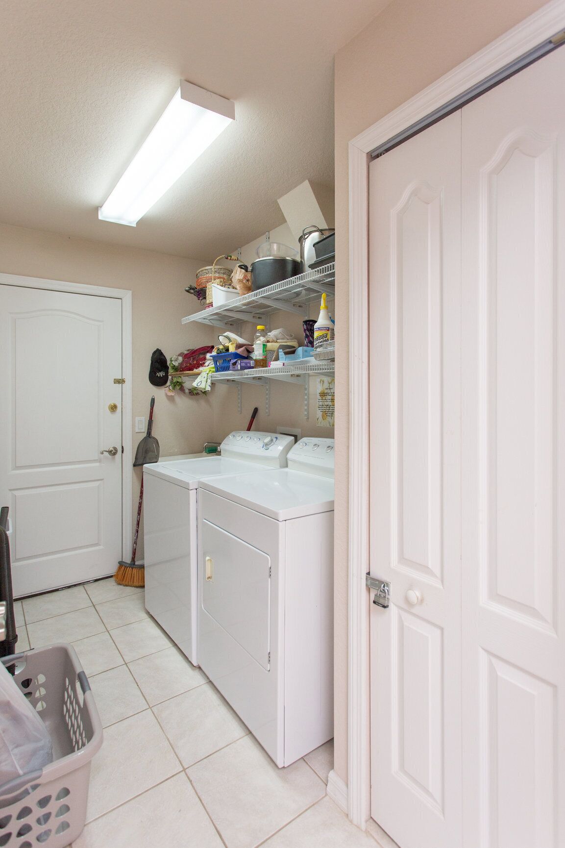 A laundry room with two white washing machines, a broom, and shelves above holding various household items.