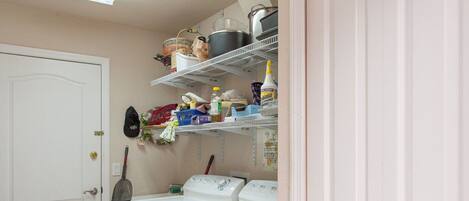 A laundry room with two white washing machines, a broom, and shelves above holding various household items.