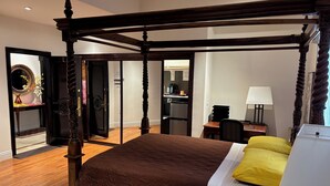 A four-poster bed with a brown quilt, a yellow pillow, and a white pillow. There is a desk with a lamp, a chair, and a mirror. A kitchen area is visible in the background.
