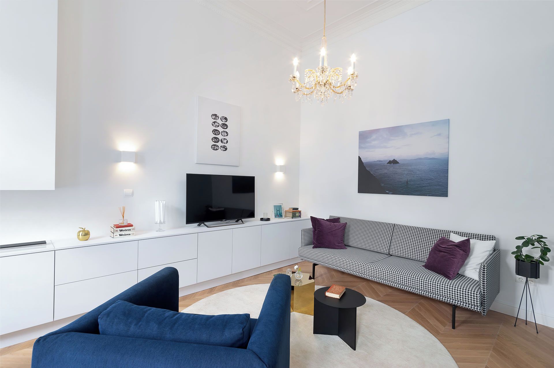 A modern living room with a sofa, a blue armchair, a TV, a chandelier, and a painting of a seascape.