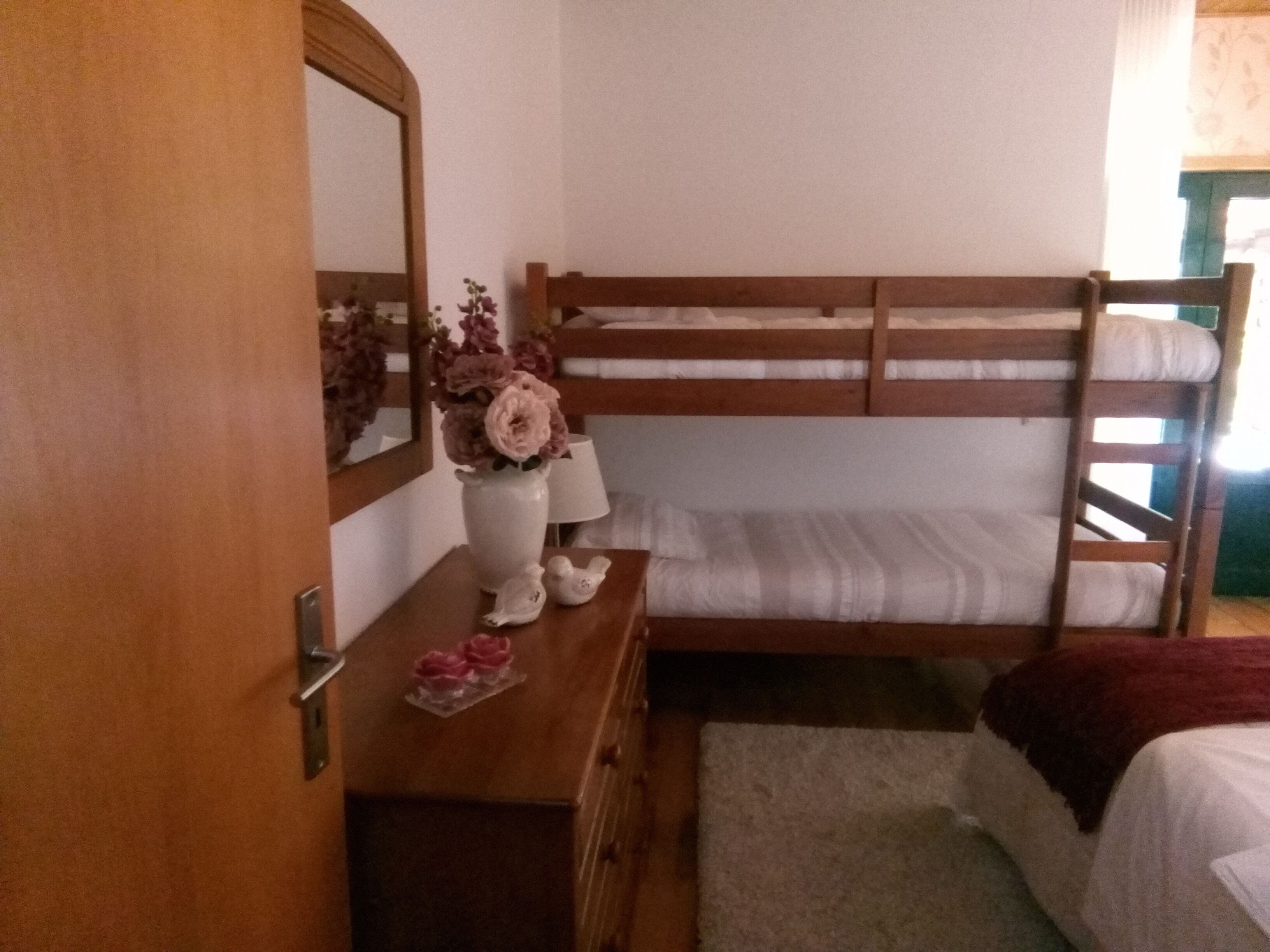 Ein Stockbett mit Holzrahmen und weißer Bettwäsche, eine Kommode mit Spiegel, eine Vase mit Blumen und eine kleine Vogelfigur.