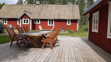 Une terrasse en bois avec une table et des chaises, un bâtiment rouge avec un toit en tuiles, et un véhicule à l’arrière-plan.