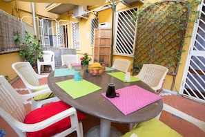 A round table with four chairs, each with a colorful cushion. There are two potted plants and a basket of fruit on the table.