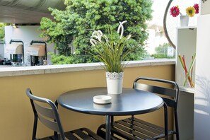 A round table with a white pot of grass, two chairs, and a window with a view of trees and buildings.