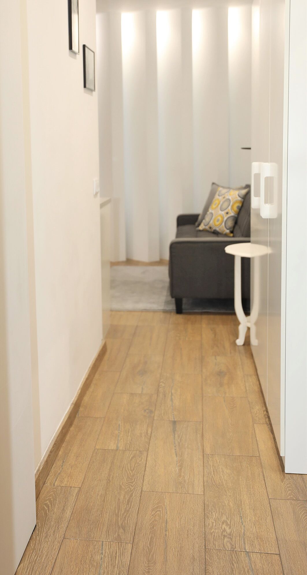 A hallway with wooden flooring, a grey sofa with patterned cushions, and a white side table.