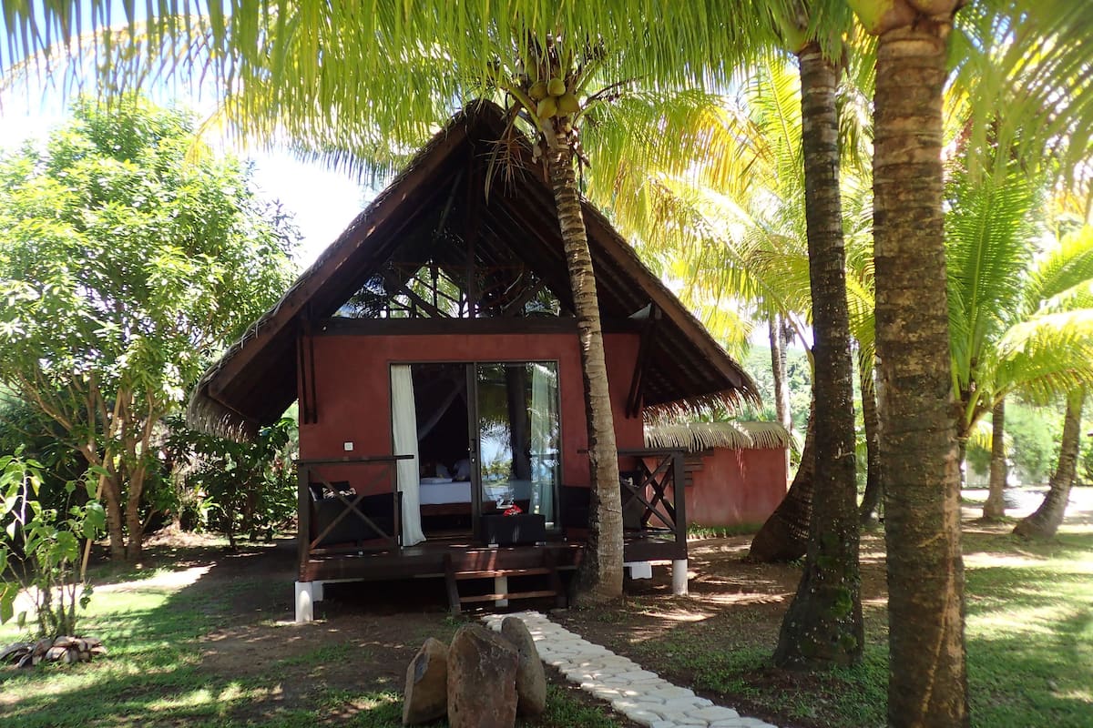 A small, single-story structure with a thatched roof, surrounded by lush greenery and palm trees.