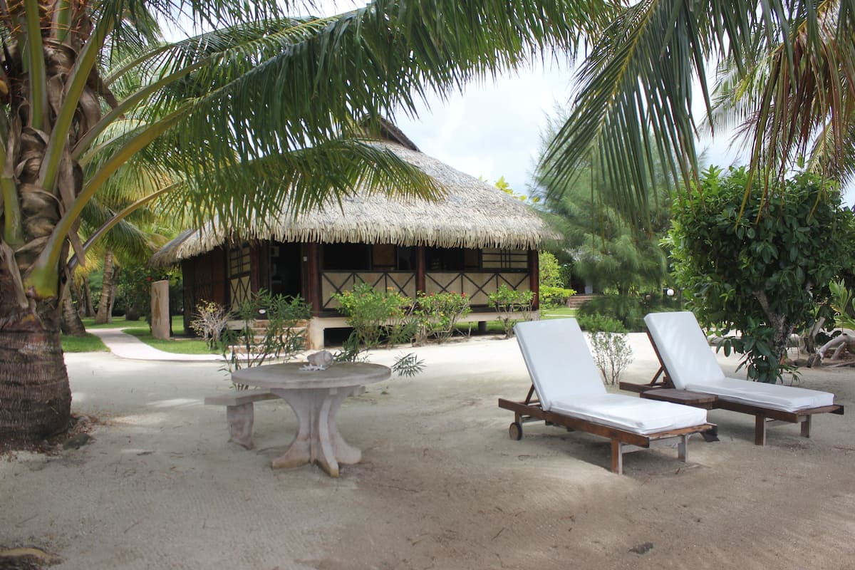 A thatched-roof hut with a sandy ground, two lounge chairs, and a round table.