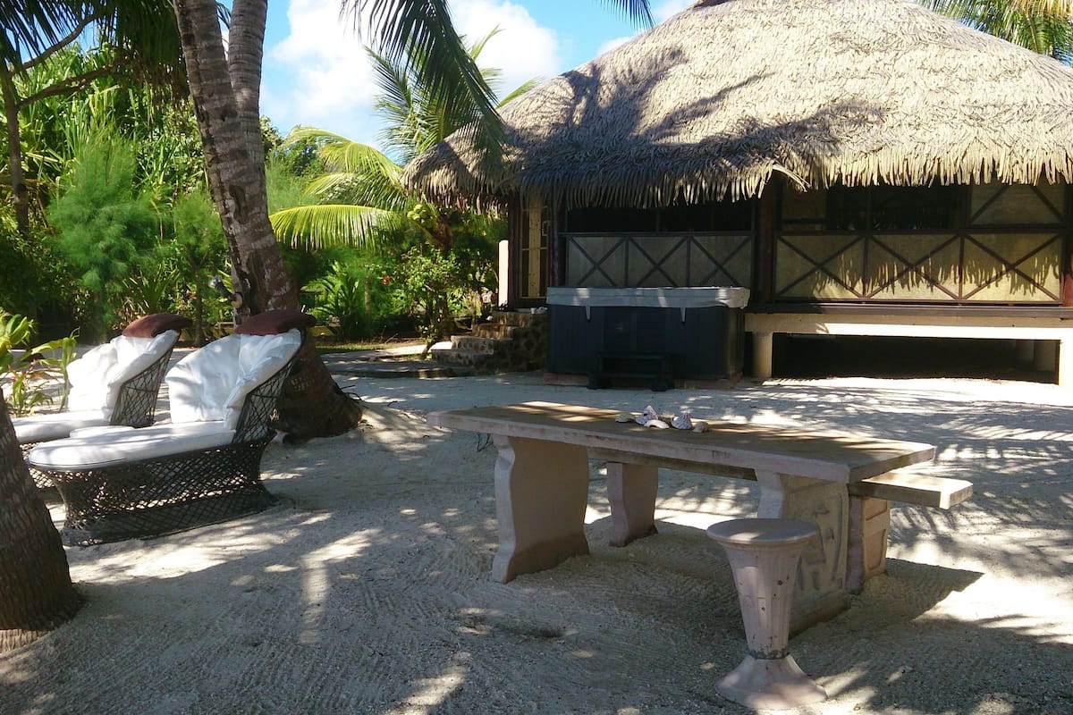 A thatched-roof hut with a bench and lounge chairs in a tropical setting.