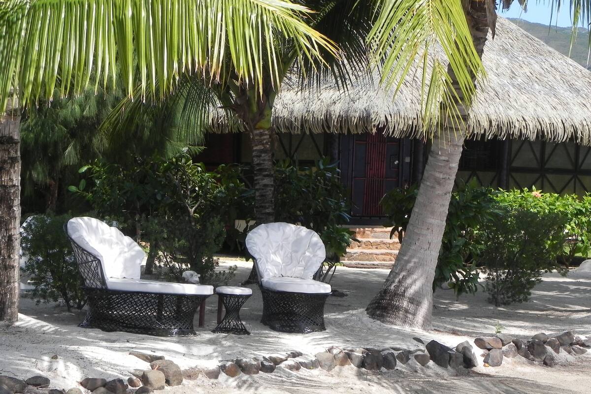 A beachside lounge with two white cushioned chairs and a small table, surrounded by palm trees and a thatched-roof hut.