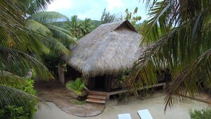 Une hutte au toit de chaume, avec un sol sablonneux, entourée de palmiers et agrémentée de deux chaises longues en bois.