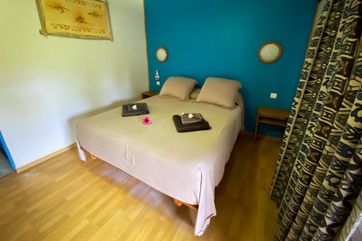 A neatly made bed with a beige comforter and pillows, a small bedside table with a lamp, and a wall-mounted clock.