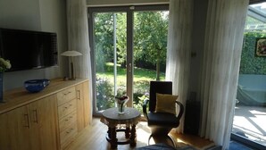 Ein Zimmer mit Holzschrank, Fernseher, rundem Tisch mit Vase, Stuhl und Blick ins Grüne durch eine Glastür.