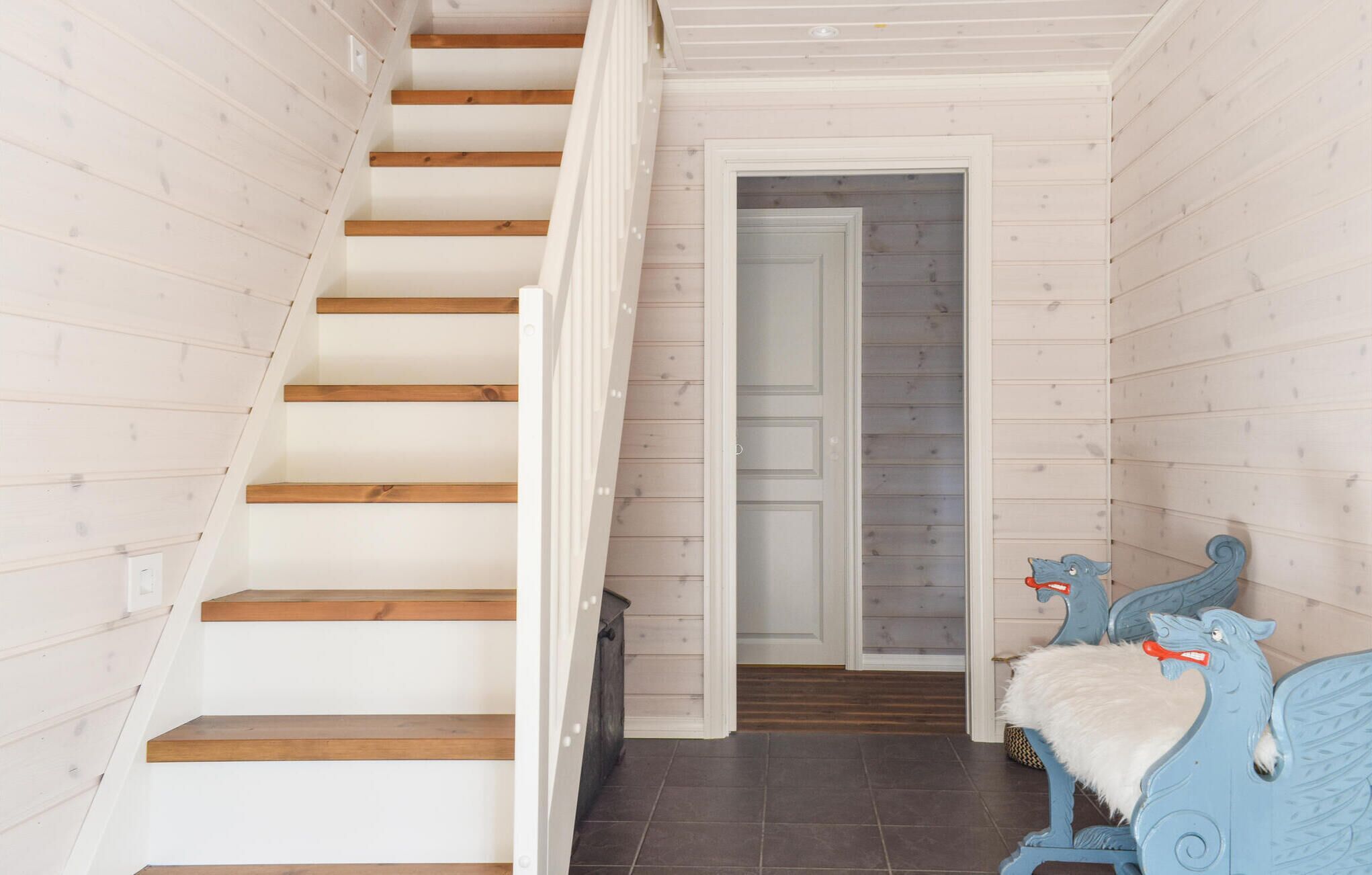 A wooden staircase with a white door at the top leading to a room with a blue chair and a white furry cushion.