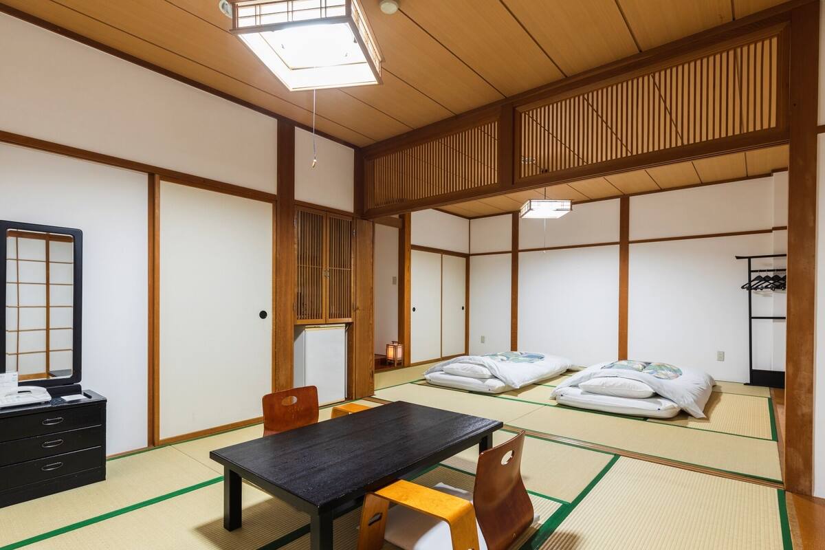 Ein traditioneller japanischer Raum mit Tatami-Boden, einem niedrigen Tisch, einem Sessel, einem Bett mit Kissen, einem Spiegel und einem Kleiderschrank.
