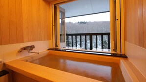 A wooden cabin bathroom with a bathtub, a window with a view of a snowy landscape, and a wooden deck outside. - Hotel Jogakura (Aomori)