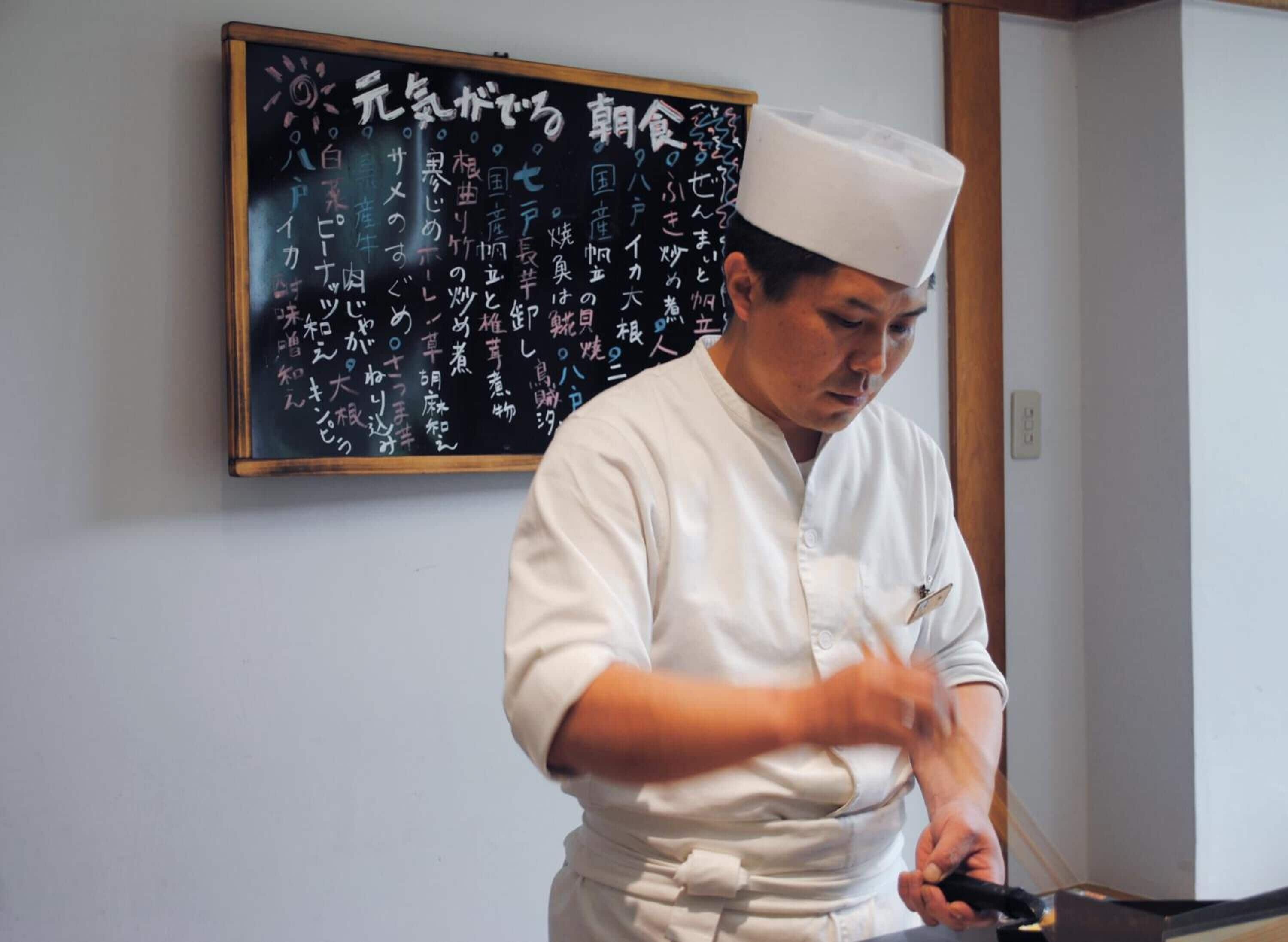 A chef in a white uniform preparing food, with a blackboard displaying Japanese text in the background.