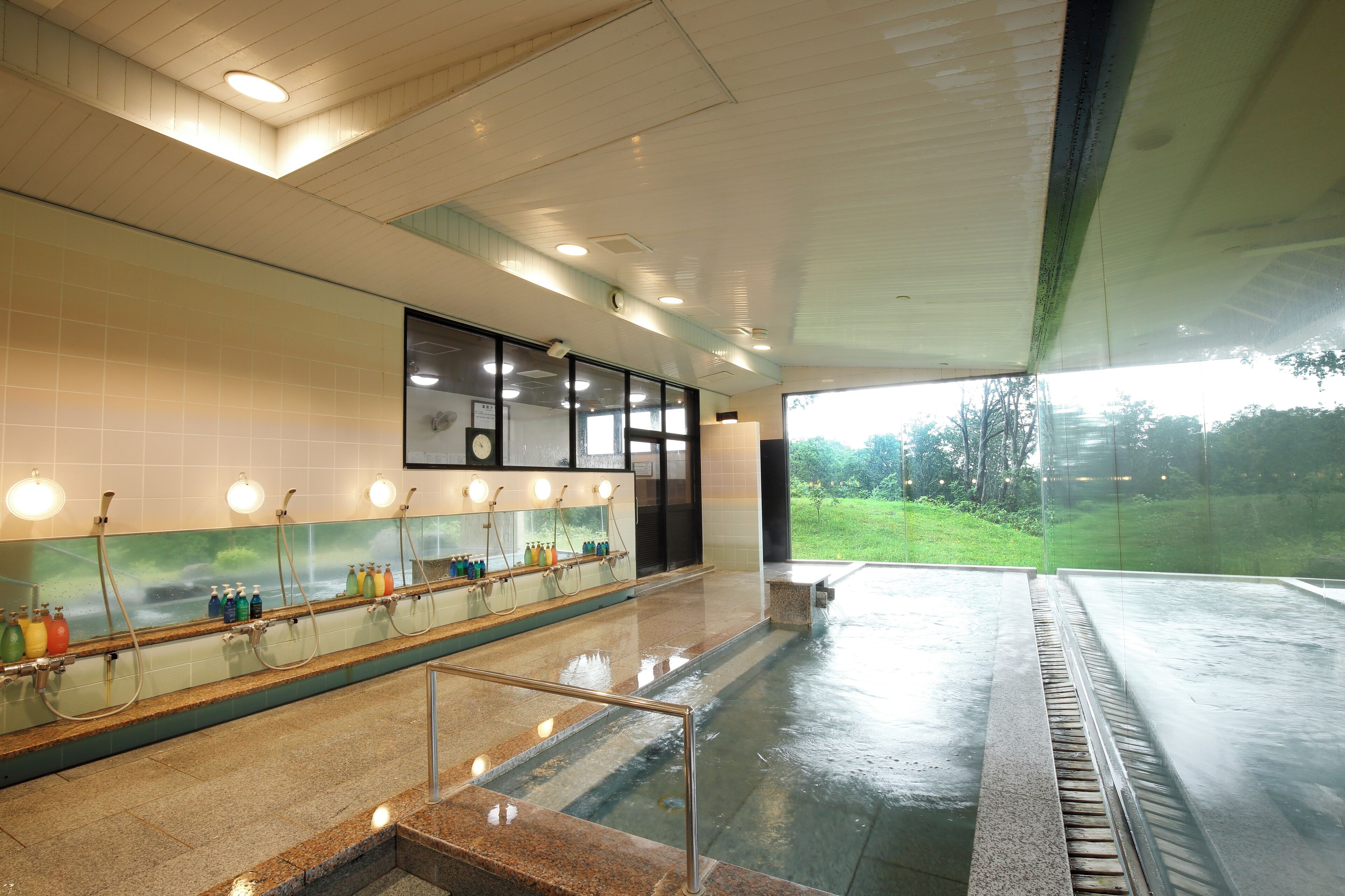 Baño público con varios lavamanos, un espejo y vistas a un paisaje verde a través de amplias ventanas.