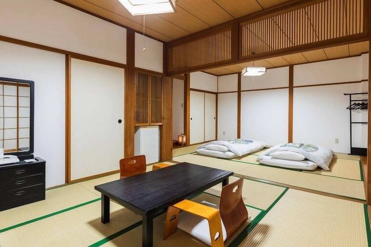 Ein traditioneller japanischer Raum mit Tatami-Böden, einem niedrigen Tisch, einem Stuhl, einer Kommode und einem Bett mit Kissen.
