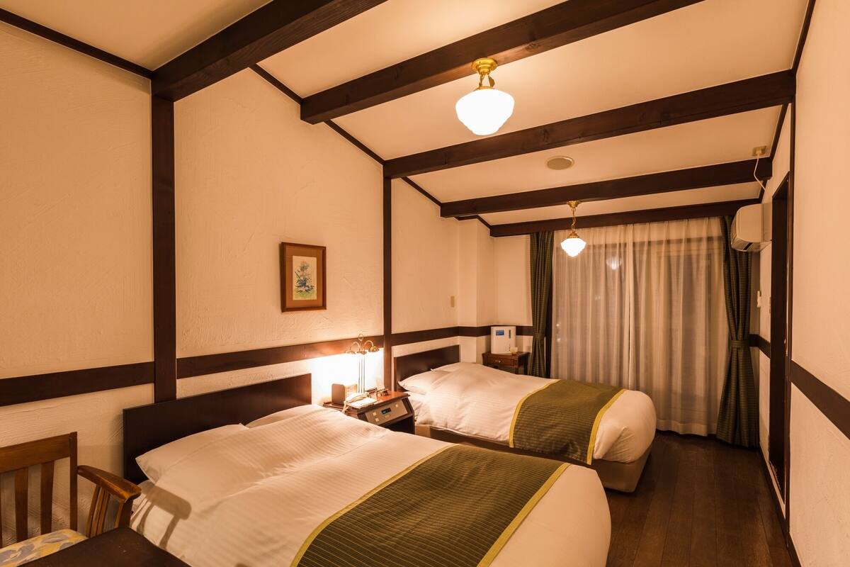 Ein Hotelzimmer mit zwei Betten, Holzbalken an der Decke, einem Nachttisch mit Lampe und einem Fenster mit Vorhängen.