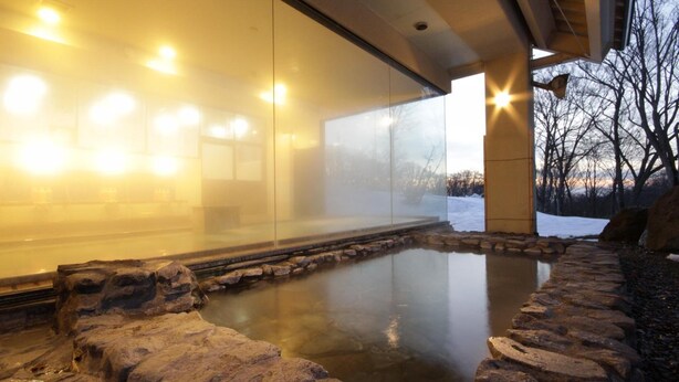 A modern spa with a glass-enclosed hot tub, stone pathway, and a view of a snowy landscape. - Hotel Jogakura (Aomori)