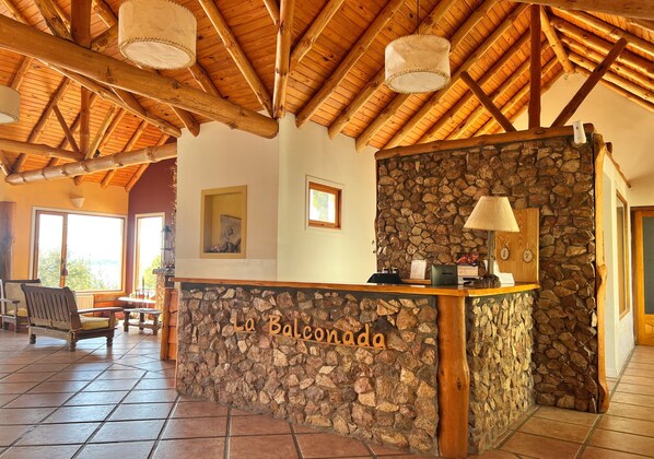 A reception area with a stone wall and wooden ceiling beams, a sign reading "La Balconada," and a view of the outdoors through large windows.
