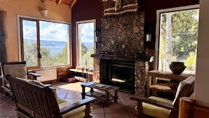 Une pièce chaleureuse avec une cheminée en pierre, des meubles en bois et de grandes fenêtres donnant sur l’extérieur.