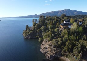 A coastal property with a house on a cliff overlooking a lake, surrounded by dense trees and mountains in the background.