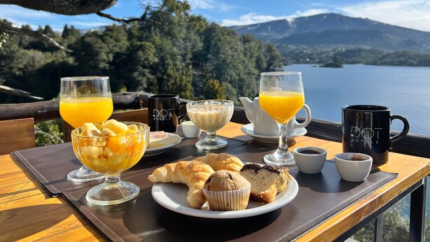 Une table de petit-déjeuner avec des croissants, des muffins et un panier de fruits, offrant une vue pittoresque sur le lac.