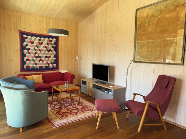 A living room with a red sofa, a blue armchair, a patterned ottoman, a wooden coffee table, a TV on a stand, and a large quilt on the wall.